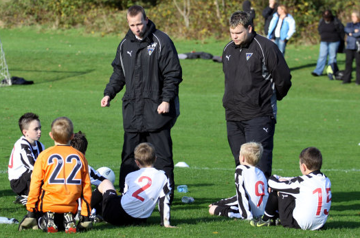 2 men standing up talking to 5 young children sitting down in football strips